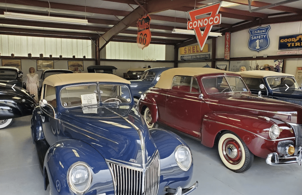 Interior of a vintage car museum featuring classic cars from the 1930s and 1940s, including a blue Ford and a maroon convertible, with vintage gas station signs like Conoco, Phillips 66, and Shell hanging from the ceiling.