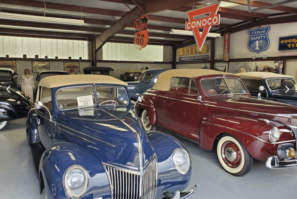 Interior of a vintage car museum featuring classic cars from the 1930s and 1940s, including a blue Ford and a maroon convertible, with vintage gas station signs like Conoco, Phillips 66, and Shell hanging from the ceiling.